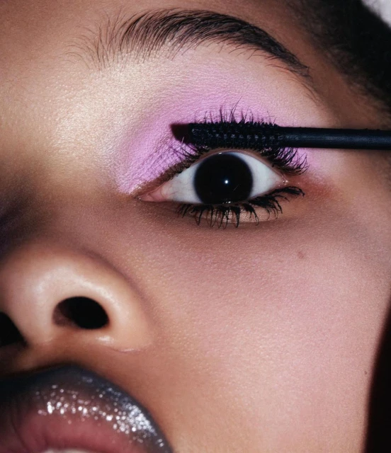 Close-up of a person's eye with lavender eye shadow and black mascara being applied with a black mascara wand.
