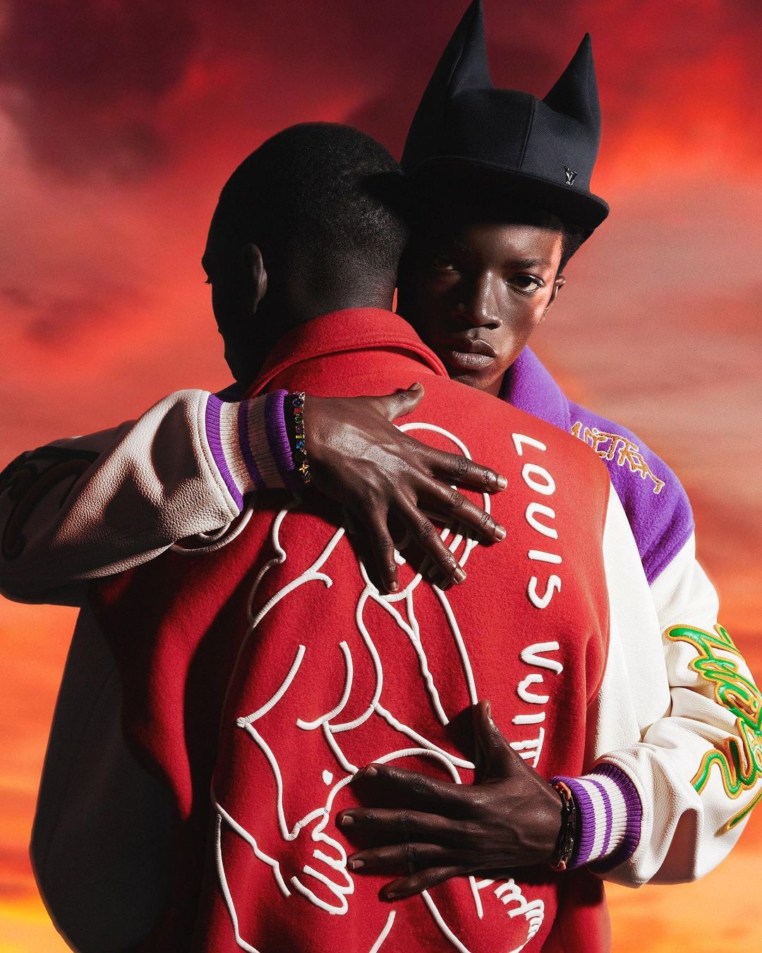 Two Black men in a close embrace. One faces forward in a black hat. The other wears a red Louis Vuitton jacket with white accents.