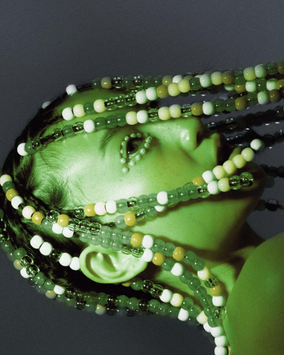 Profile of an adult woman with beaded braids and beaded decoration around the eye, set against a dark green-toned background.