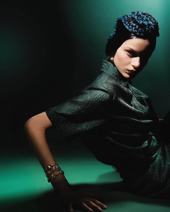 A young fair-skinned model in dark teal textured top and headwear, strikes a dramatic pose with bracelet against a dark background.