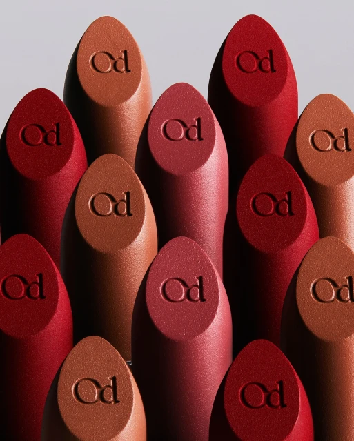 Close-up of multiple lipstick bullets in varying shades of red and brown with the brand logo embossed on each, against a grey background.