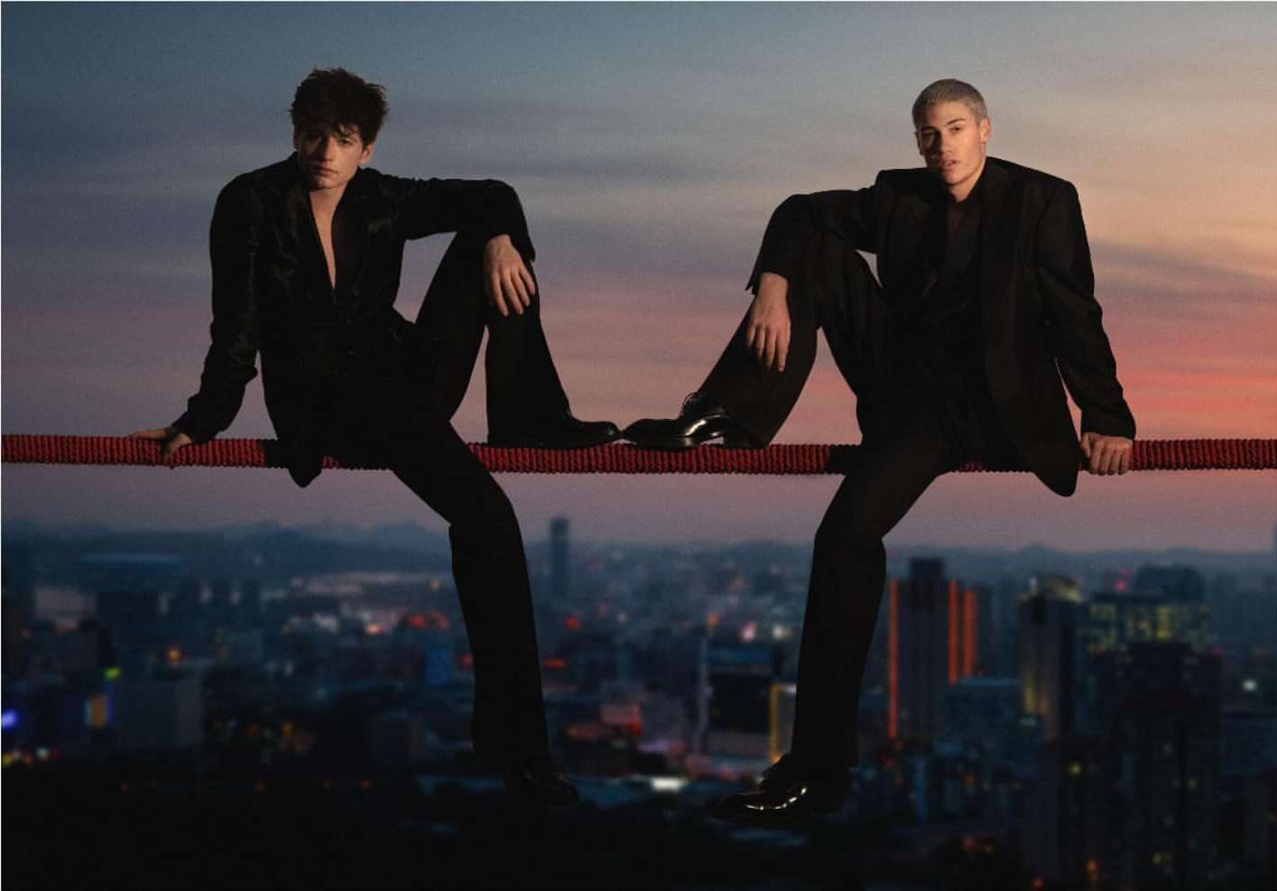 Two young men in black suits sit on a red rope suspended over a nighttime cityscape silhouetted against a colorful sunset sky backdrop.