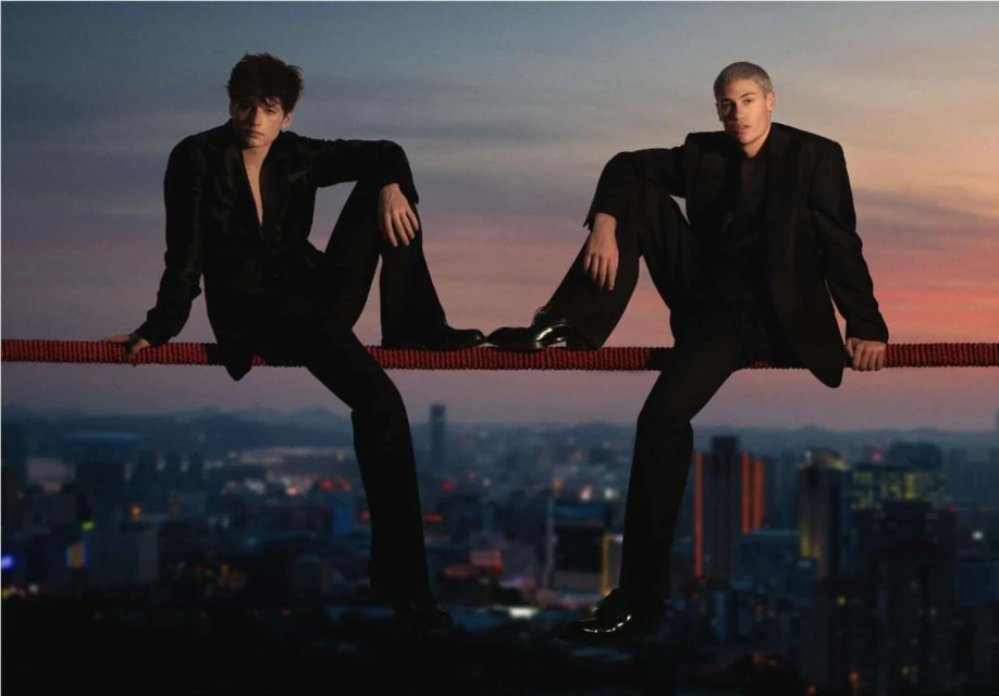 Two young men in black suits sit on a red rope suspended over a nighttime cityscape silhouetted against a colorful sunset sky backdrop.