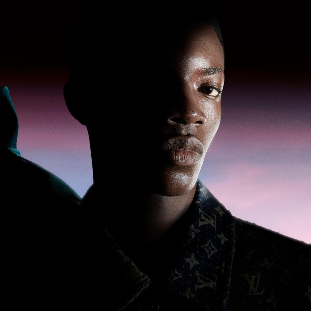Close-up studio portrait of a Black male model wearing a jacket with logos; half of the model's face is obscured by shadow.