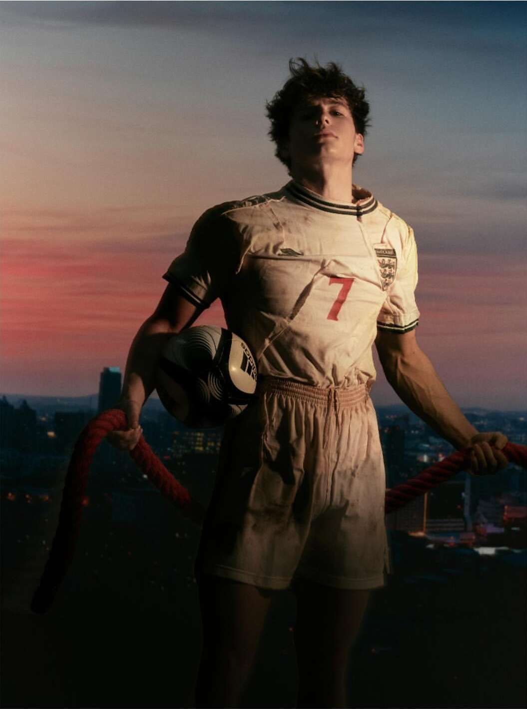 Male soccer player in uniform with a soccer ball stands against a backdrop of a sunset and cityscape, athletic and dramatic.
