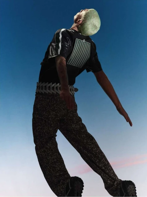 Low angle view of a slim adult male with light skin wearing a black t-shirt, patterned pants, and a decorative belt against a blue sky.