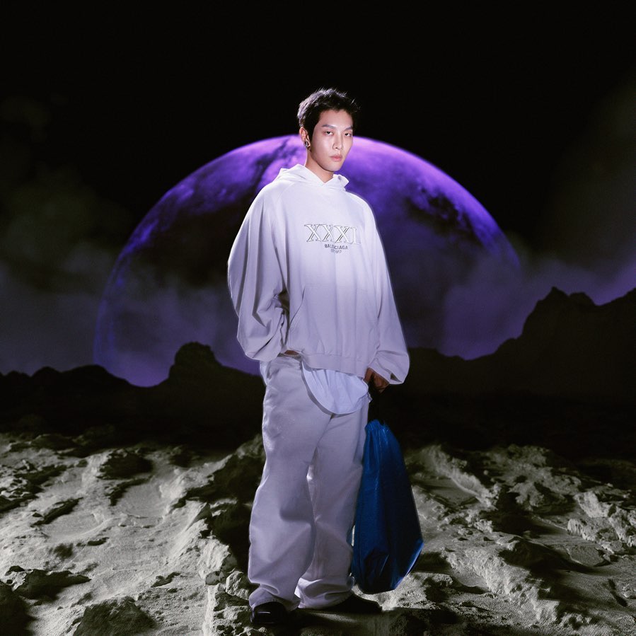 Asian man poses in grey hoodie and pants with blue bag, standing on moon-like surface in front of a large planet against a black backdrop.