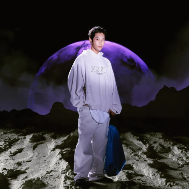 Asian man poses in grey hoodie and pants with blue bag, standing on moon-like surface in front of a large planet against a black backdrop.