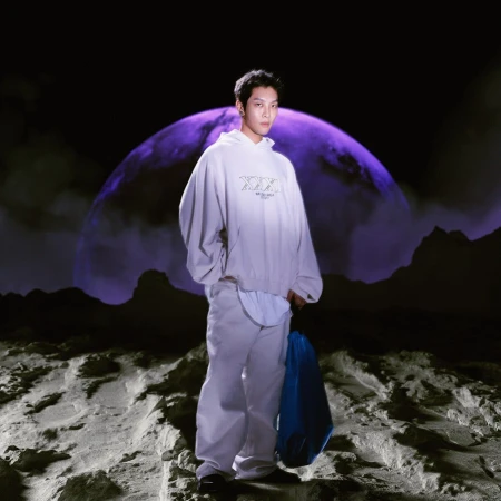 Asian man poses in grey hoodie and pants with blue bag, standing on moon-like surface in front of a large planet against a black backdrop.