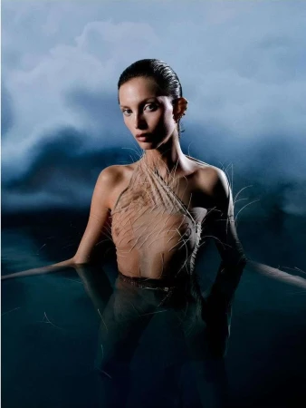 Model with slicked-back wet hair in a textured feathered garment standing in water against a cloudy sky background
