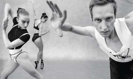 Monochrome image of a male dancer in the foreground with outstretched hand and two female dancers posing in the background.