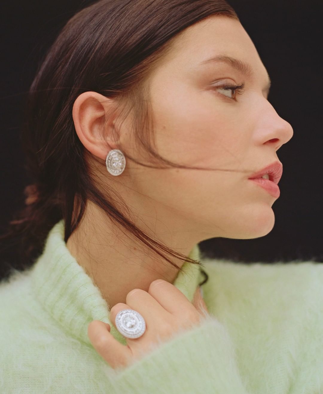 Adult female in a green sweater wears an oval earring and ring with silver stones, posing against a black background in studio lighting.