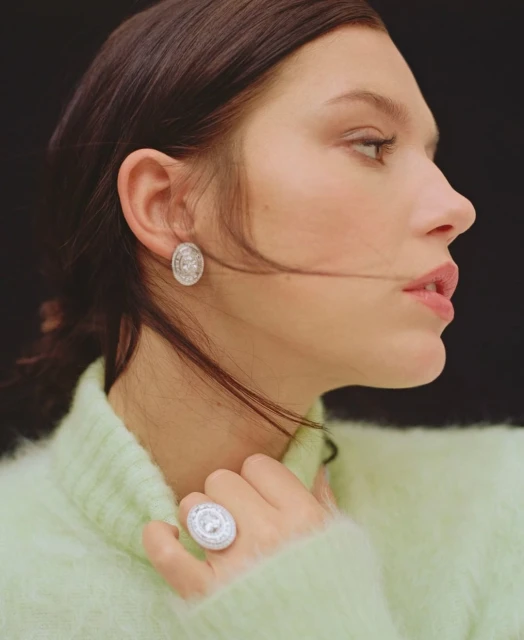 Adult female in a green sweater wears an oval earring and ring with silver stones, posing against a black background in studio lighting.