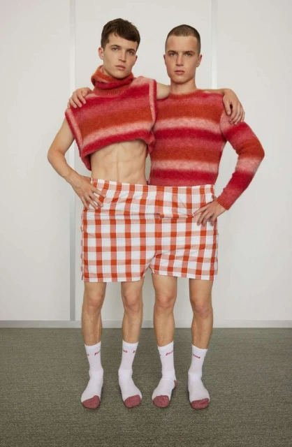 Two male models pose indoors wearing matching red and orange striped sweaters and red and white checkered shorts with white socks and sneakers.