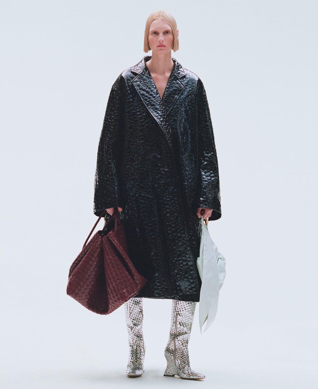 Full-length shot of a female fashion model in a black vinyl trench coat, silver snake-skin boots, holding a burgundy woven tote bag.