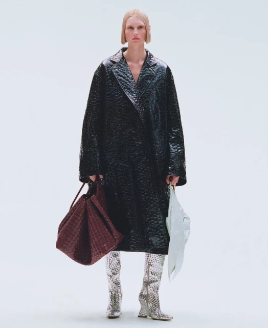 Full-length shot of a female fashion model in a black vinyl trench coat, silver snake-skin boots, holding a burgundy woven tote bag.