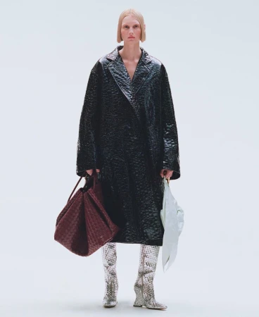 Full-length shot of a female fashion model in a black vinyl trench coat, silver snake-skin boots, holding a burgundy woven tote bag.