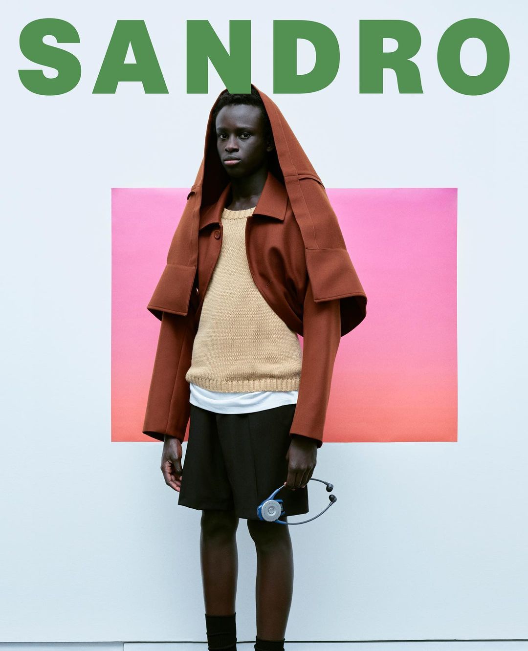 Fashionable adult male model showcases Sandro apparel in a studio shot, posing with a stethoscope, set against a gradient backdrop.