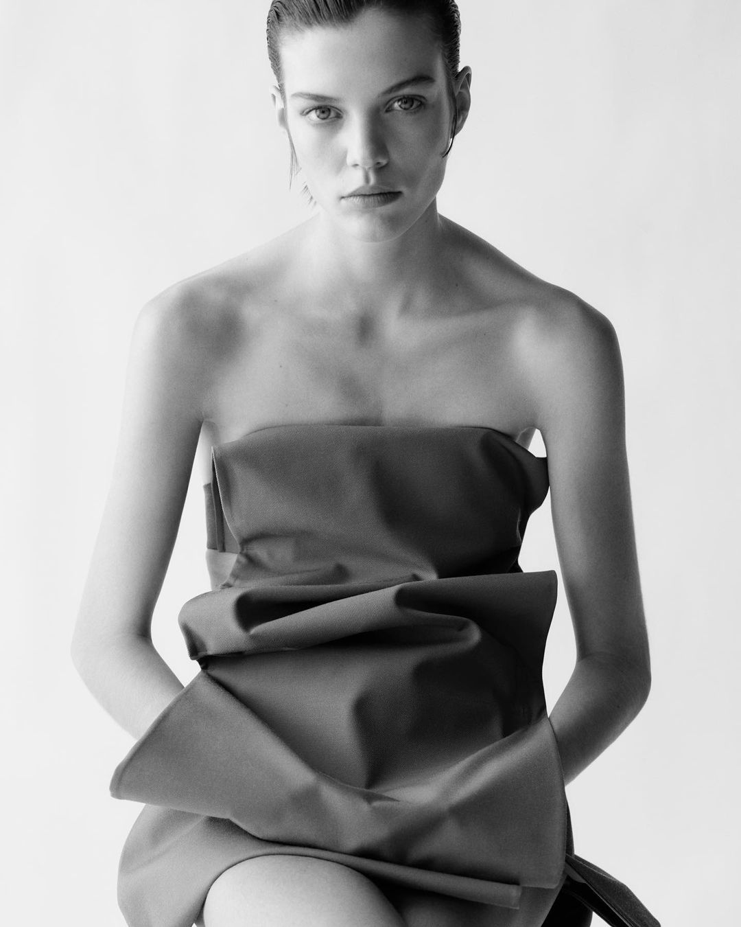 Black and white portrait of a young fair-skinned woman in a strapless top with a large bow, with slicked-back hair, looking at the camera.