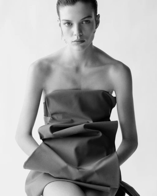 Black and white portrait of a young fair-skinned woman in a strapless top with a large bow, with slicked-back hair, looking at the camera.