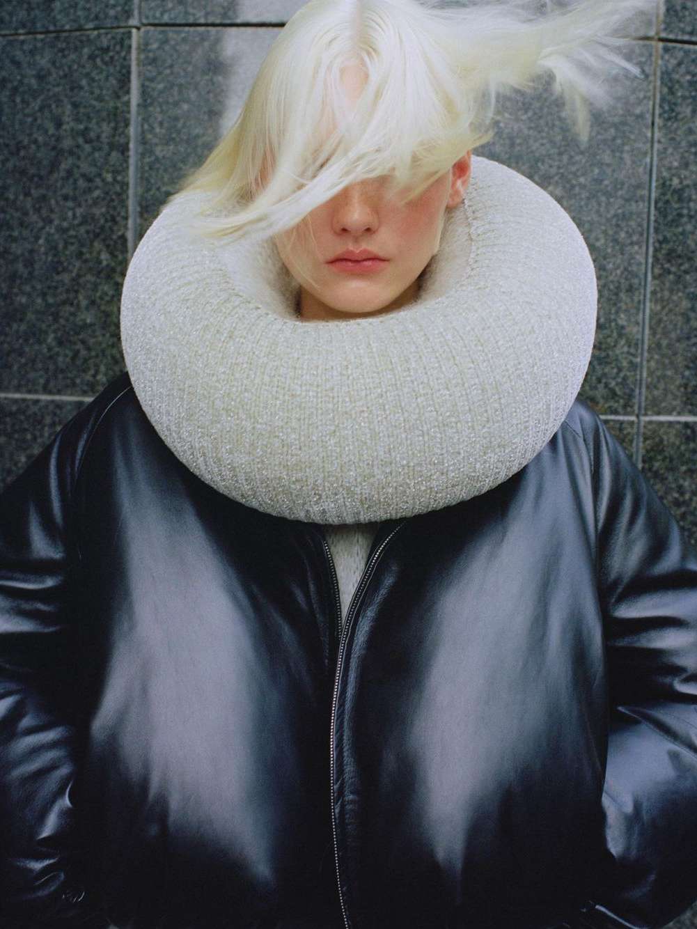 Fashion shot of a person with dyed blonde hair wearing a black jacket and a white knit collar in front of a grey tiled wall.