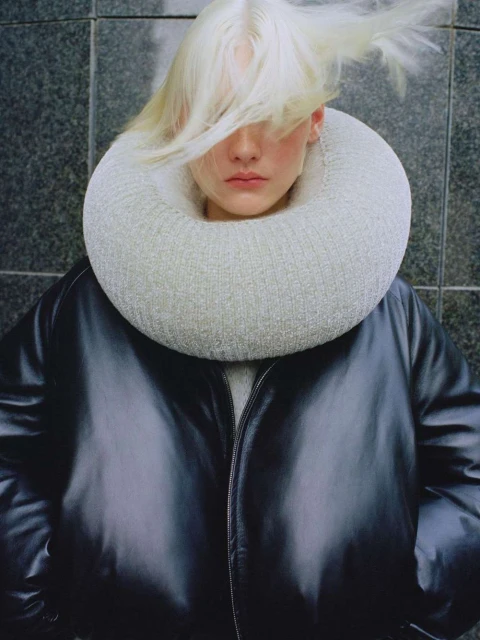 Fashion shot of a person with dyed blonde hair wearing a black jacket and a white knit collar in front of a grey tiled wall.