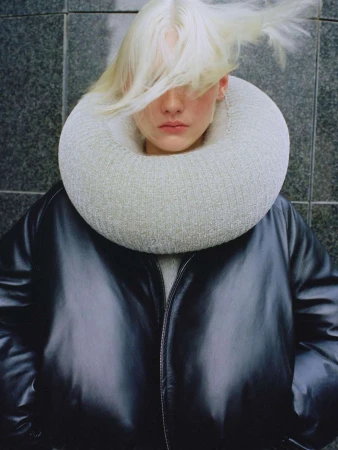 Fashion shot of a person with dyed blonde hair wearing a black jacket and a white knit collar in front of a grey tiled wall.