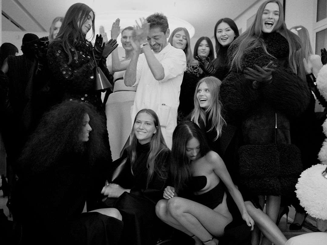 Black and white shot of a group of fashion models and a designer backstage at a runway show posing together in a photo studio.