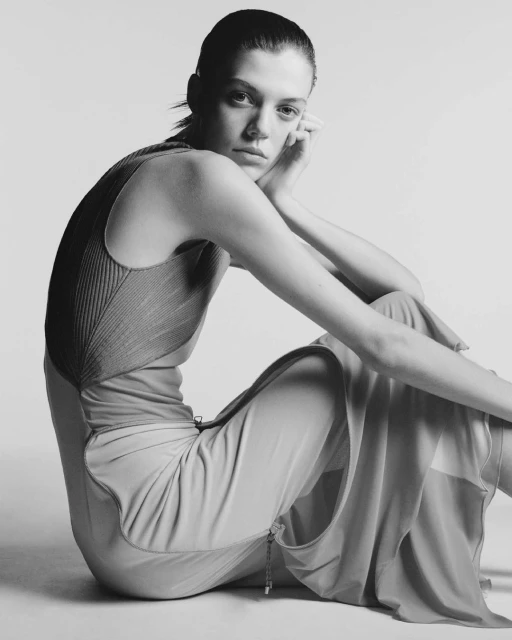 Monochrome portrait of a young adult woman in a dress, sitting with her head resting on her hand in a photo studio setting.