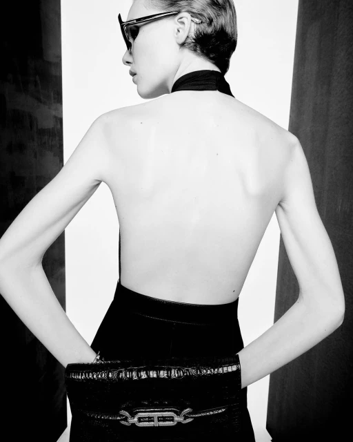Woman with dark sunglasses and bare back wearing a black outfit and holding a black clutch purse in a high contrast studio setting.