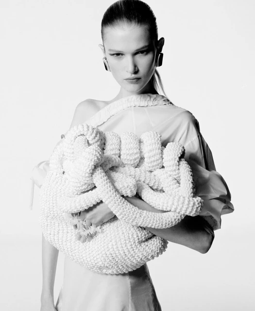 Black and white studio shot of a model holding a large, intricately knotted white bag, wearing a simple dress and statement earrings.