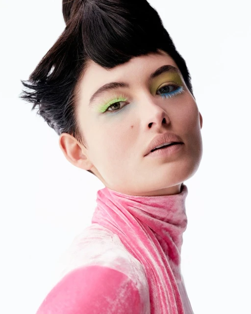 Close-up beauty shot of a woman with black hair and vivid makeup featuring colorful eyeshadow and pink velvet turtleneck top on a white background.