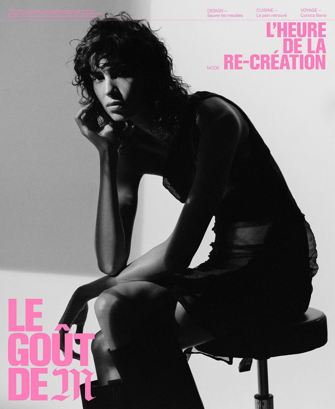Brunette woman in fashion dress and boots sitting on stool with magazine cover featuring bold pink text in a photo studio.
