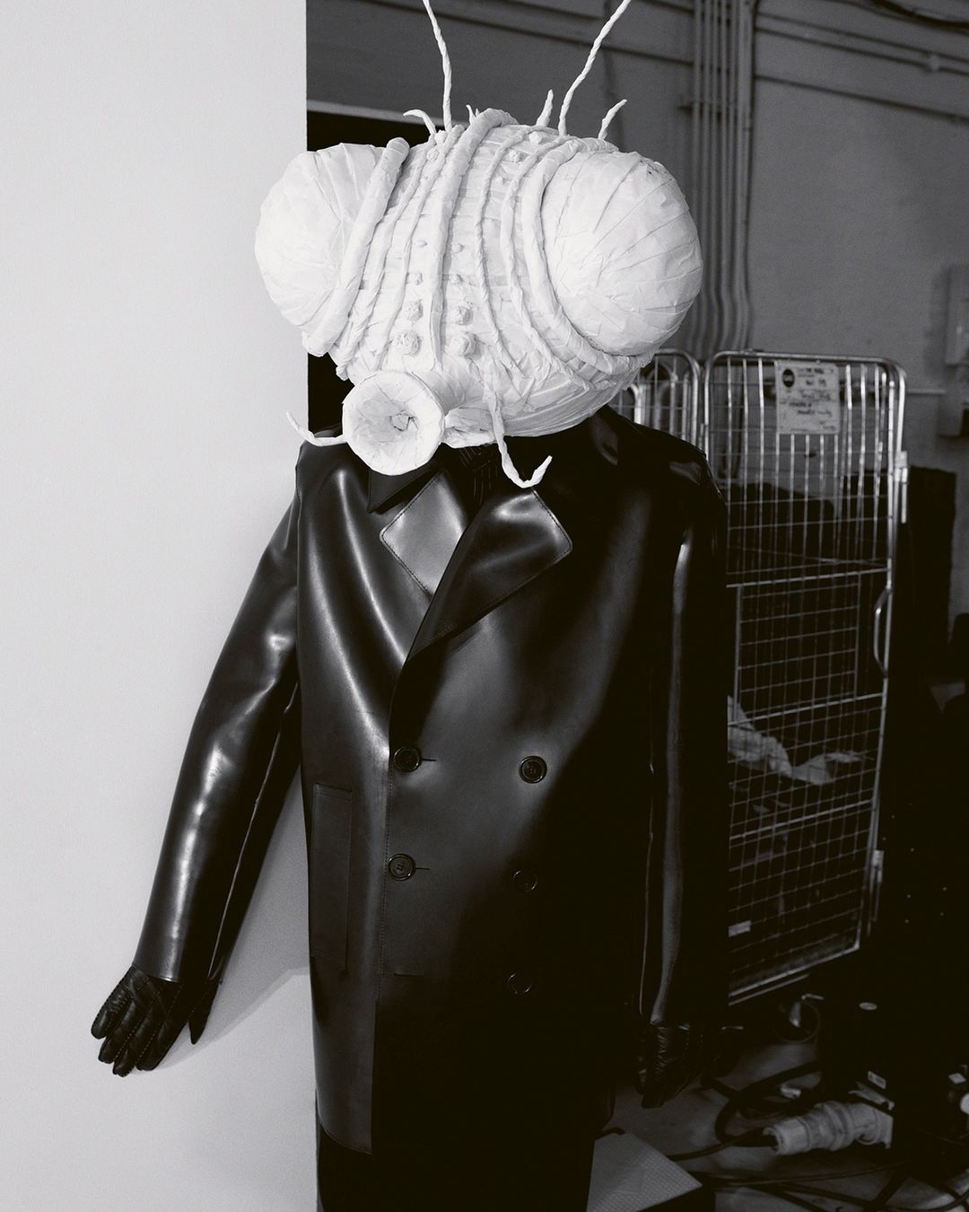 Black and white image of a mannequin wearing a black double-breasted coat with a large insect head in a studio setting.