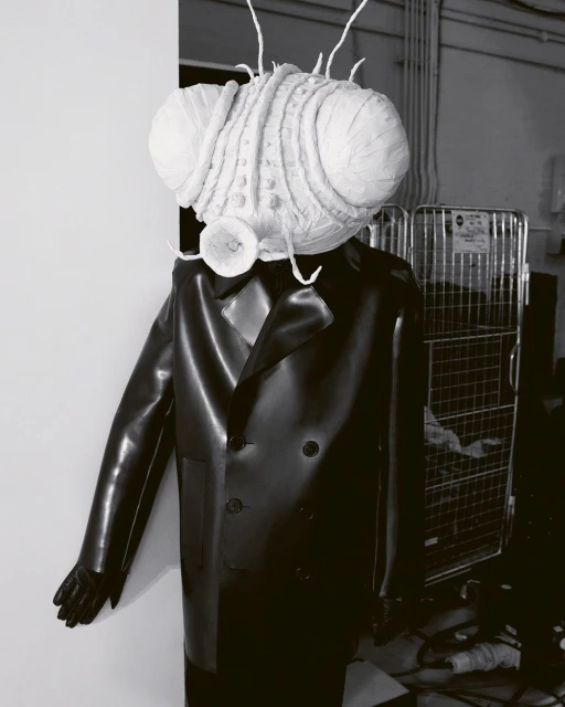 Black and white image of a mannequin wearing a black double-breasted coat with a large insect head in a studio setting.