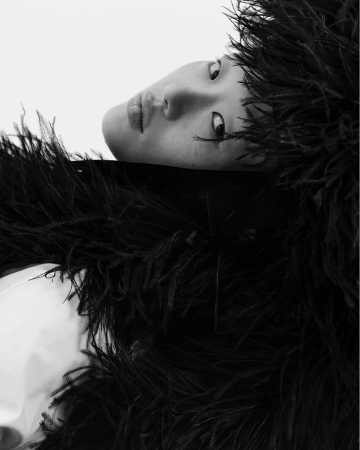 Close-up black-and-white portrait of a model with dramatic eye makeup, posing in dark feathered shawl against a bright, neutral backdrop.