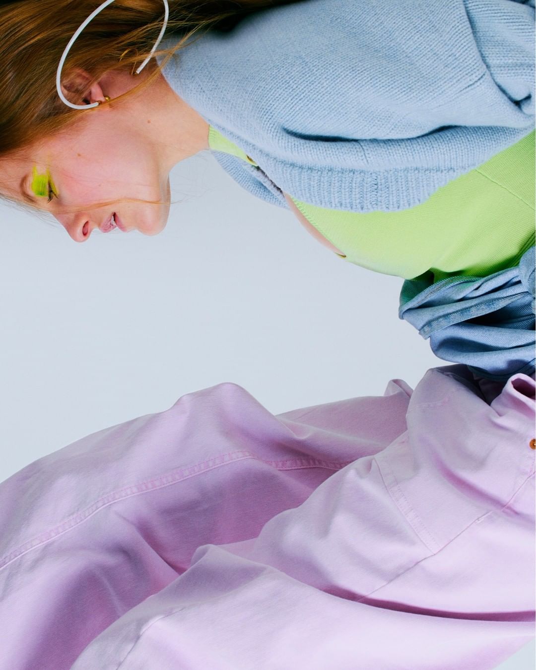 Woman with lime green eyeshadow, light blue jacket, and lilac pants in a studio setting, shot at an upward angle.