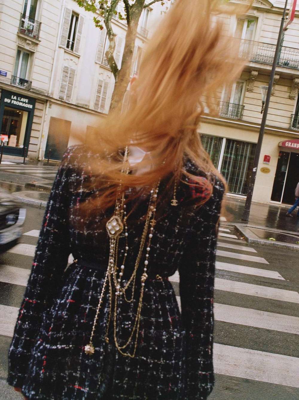 Adult woman with flowing red hair wears a Chanel tweed jacket and layered pearl and gold necklaces on a city street.