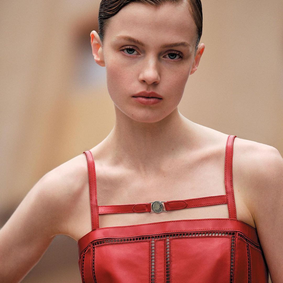 Close-up of a fair-skinned adult woman with slicked-back short brown hair, wearing a red leather dress, against a tan blurred background.