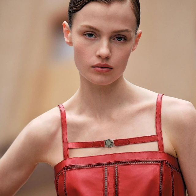 Close-up of a fair-skinned adult woman with slicked-back short brown hair, wearing a red leather dress, against a tan blurred background.