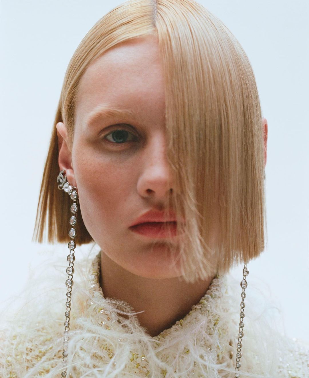 Close-up portrait of a fair-skinned adult woman with blonde, straight hair, wearing dangling earrings and a feather-trimmed jacket, indoors.