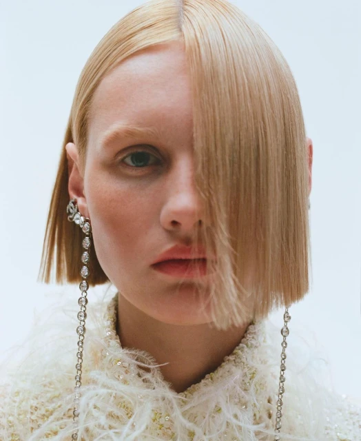 Close-up portrait of a fair-skinned adult woman with blonde, straight hair, wearing dangling earrings and a feather-trimmed jacket, indoors.