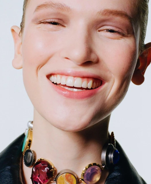 Close-up of a smiling adult woman wearing a multicolored gemstone statement necklace and a black leather jacket against a white backdrop.