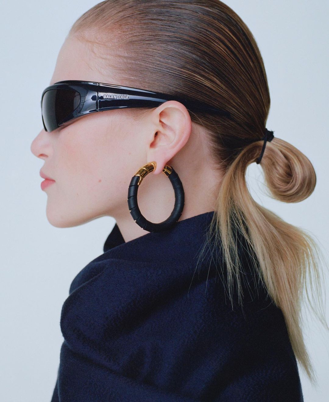 Profile of fair-skinned woman with sunglasses, hoop earrings, and navy jacket against a neutral backdrop in a portrait style.