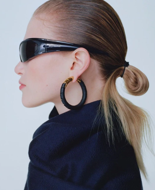 Profile of fair-skinned woman with sunglasses, hoop earrings, and navy jacket against a neutral backdrop in a portrait style.
