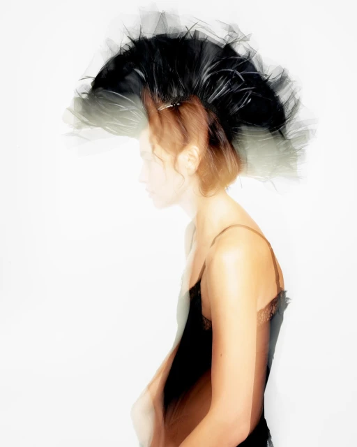 Profile view of a young woman with a decorative black feathered headpiece, wearing a black camisole against a white background in a studio shot.