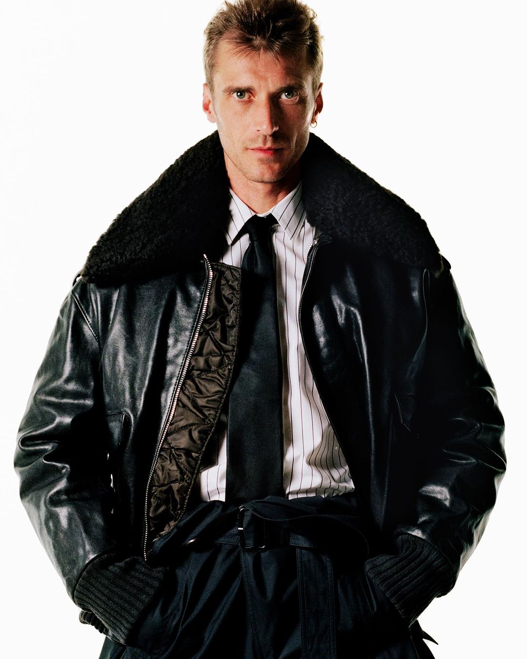 Man wearing a dark leather bomber jacket with a shearling collar over a striped shirt and tie against a plain white background