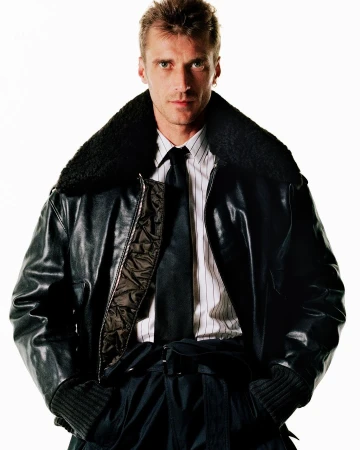 Man wearing a dark leather bomber jacket with a shearling collar over a striped shirt and tie against a plain white background