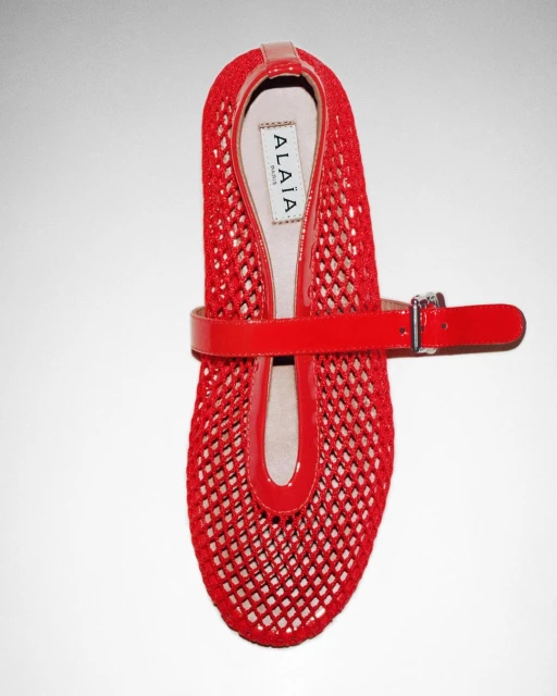 Red Alaïa ballet flat shoe with patent leather strap, buckle closure and mesh upper on white background, fashionable and stylish.