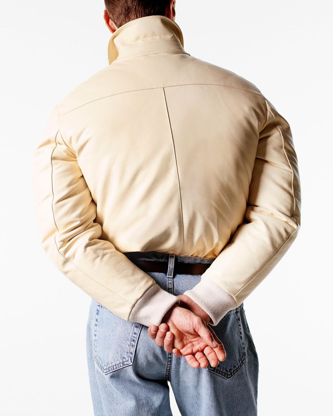 Back view of a person wearing a cream-colored jacket and blue jeans against a white background, showcasing fashion apparel.